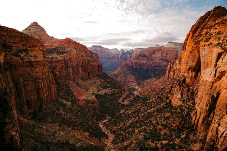 A dramatic, high-angle view looking down into a steep, narrow canyon framed by towering, reddish-orange sandstone cliffs, with a winding road and sparse green vegetation visible on the canyon floor below a cloudy sky.