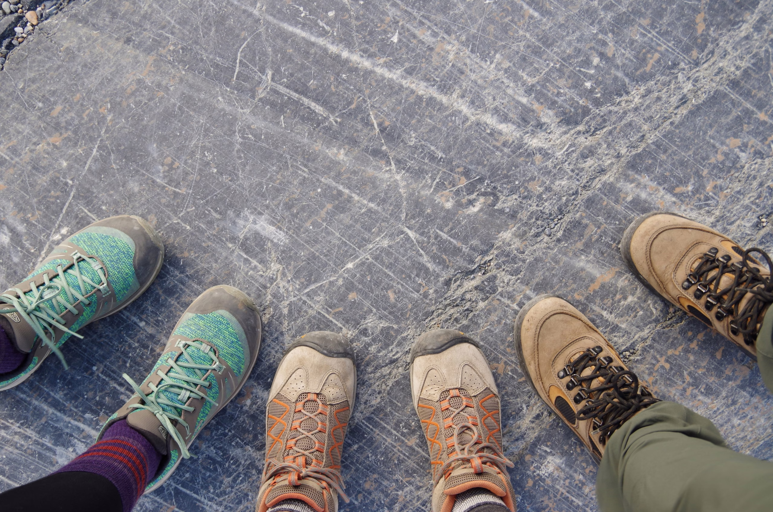 The Ultimate Outdoor Footwear Guide (All Types Explained & More) Four pairs of hiking boots stand on a textured gray stone surface. The boots are in varying shades of green, orange, and brown, conveying adventure.