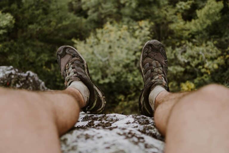 Hiker's legs in brown hiking shoes overlook a forested valley from a rocky ledge. The scene conveys a sense of adventure and tranquility.