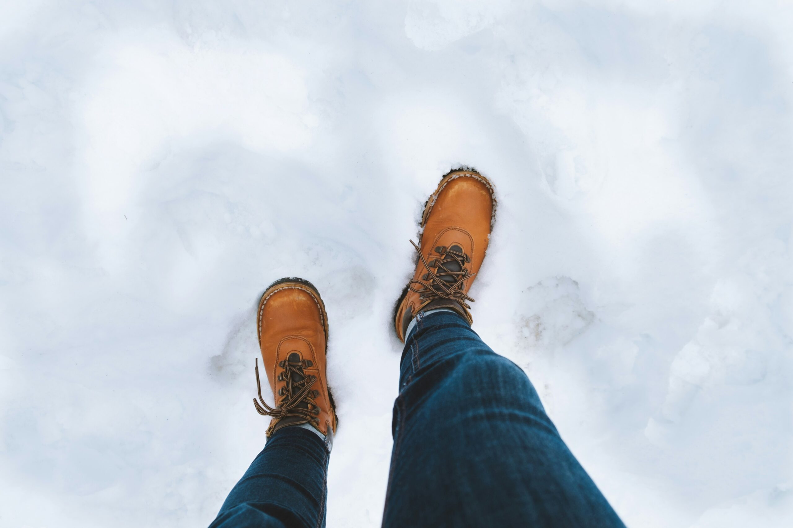 7 Best Snowshoes For Winter Hiking & Mountaineering Brown boots stand in fresh snow, surrounded by footprints. The dark jeans create a contrast against the bright white snow, evoking a wintery feel.