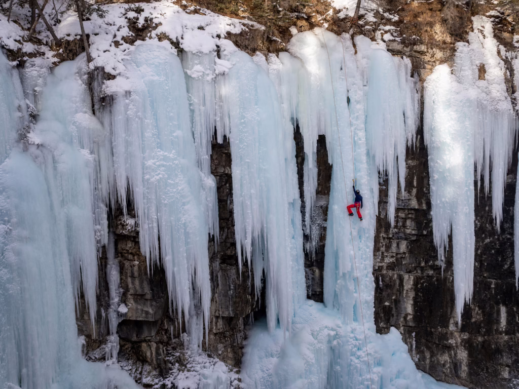 Mountaineering Beginners Guide: Preparing Your First Ascent A person in red pants and a dark jacket is ice climbing a massive frozen waterfall. The icy formations are draped over a rocky cliff, surrounded by snow.