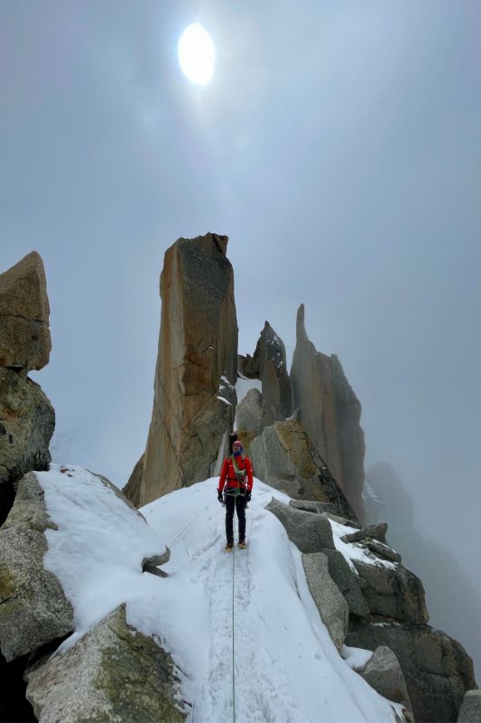 Mountaineering Beginners Guide: Preparing Your First Ascent A climber reaches a snowy mountain summit, with a prominent rock formation towering in the background.