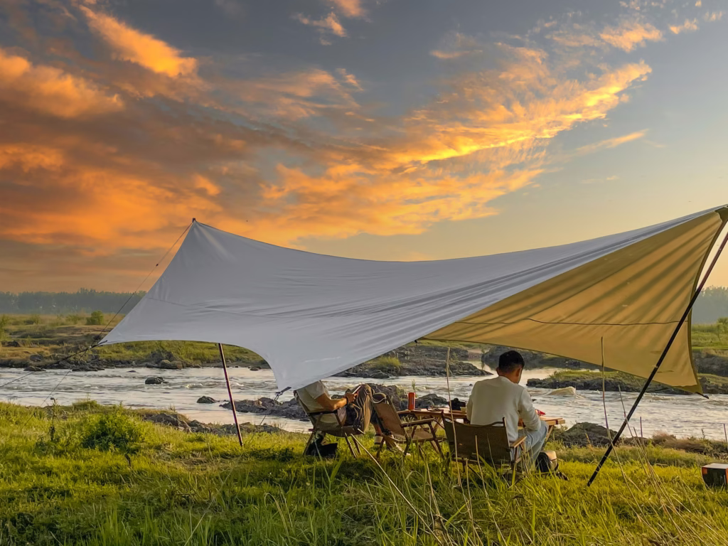 A person sits under a large white tarp by a river, surrounded by grass. The sky is vibrant with a dramatic orange sunset, creating a tranquil atmosphere.