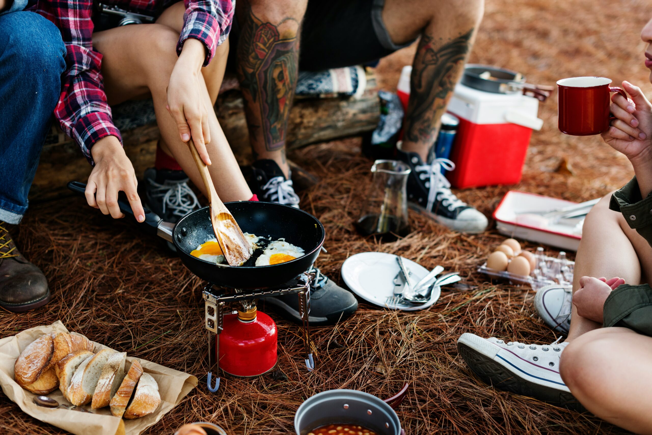 Multi-Day Hiking Nutrition: Expert Tips by Nancy Clark People cooking breakfast outdoors with a portable stove on pine needles. Frying eggs, sliced bread, coffee mugs, eggs, and a cooler indicate a relaxed camping setting.