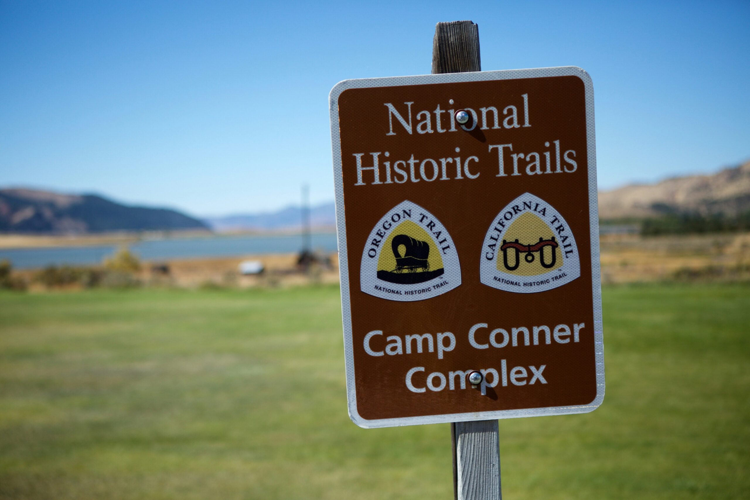 How to Turn Wild Camping Into a Glamping Experience Brown sign for "National Historic Trails" at Camp Conner Complex, featuring Oregon and California Trail emblems. Scenic view of lake and mountains in background.