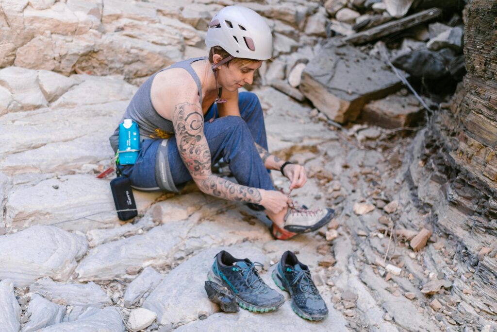 10 Best Approach Shoes For Hiking & Climbing (2026) Tattooed climber sitting on rocky ground, wearing a helmet, tying climbing shoes. Hiking shoes and climbing gear nearby. Focused, determined expression.