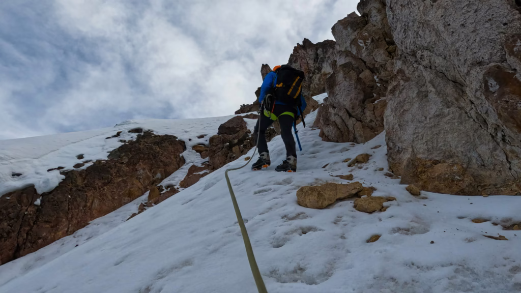 Mountaineering Beginners Guide: Preparing Your First Ascent A climber in winter gear ascends a snowy, rock-strewn slope, secured by a rope. The scene is set against a backdrop of cloudy sky, conveying adventure.