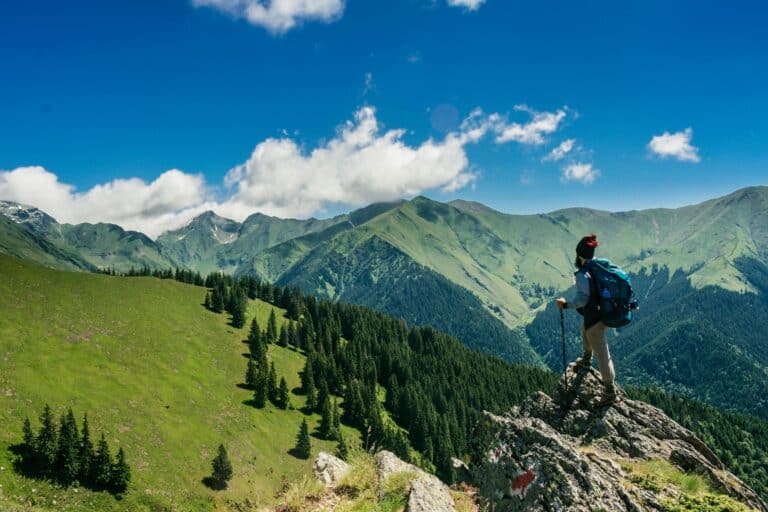 Hiker with a blue backpack stands on a rocky outcrop, overlooking lush green mountains under a clear blue sky, conveying a sense of adventure and awe.