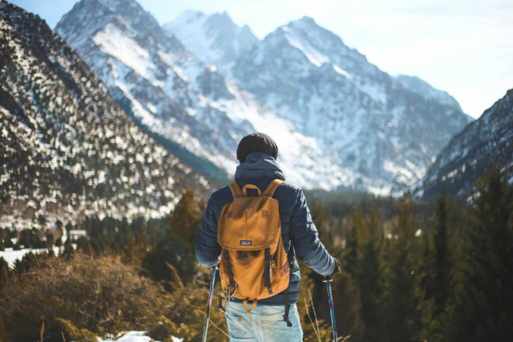 Hiker in an insulated jacket with a yellow pack and trekking poles, looking up a forested valley toward snow-covered peaks