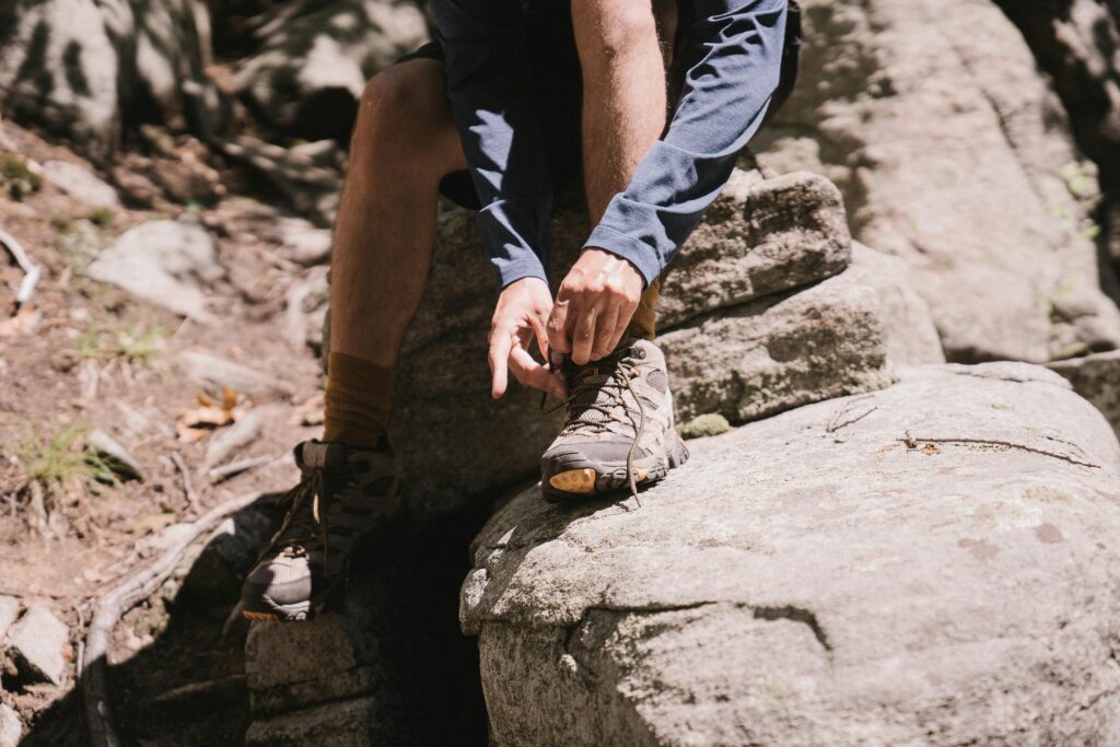 10 Best Approach Shoes For Hiking & Climbing (2026) Person tying hiking boots while sitting on a large rock in a sunlit forest. They wear a blue long-sleeve shirt and shorts, conveying an adventurous mood.