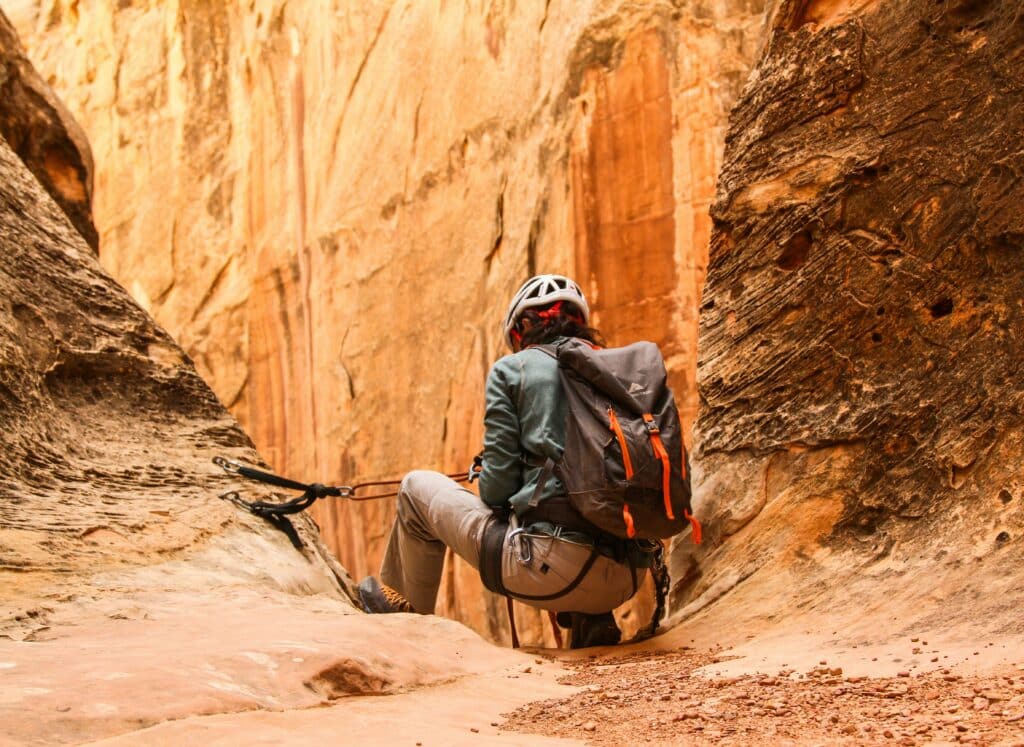 Hiker with a large backpack and a helmet lowering into a narrow sandstone canyon