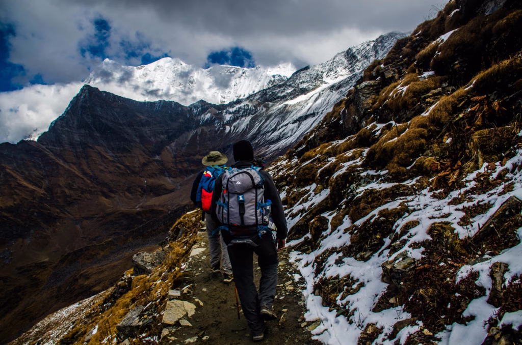 Mountaineering Beginners Guide: Preparing Your First Ascent Two hikers with backpacks walk along a snowy mountain trail under a cloudy sky. Majestic peaks loom in the distance, creating a sense of adventure.