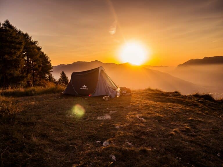 A lone tent on a grassy hilltop basks in the warm glow of a sunset. Mountains are silhouetted in the background, creating a serene and peaceful atmosphere.