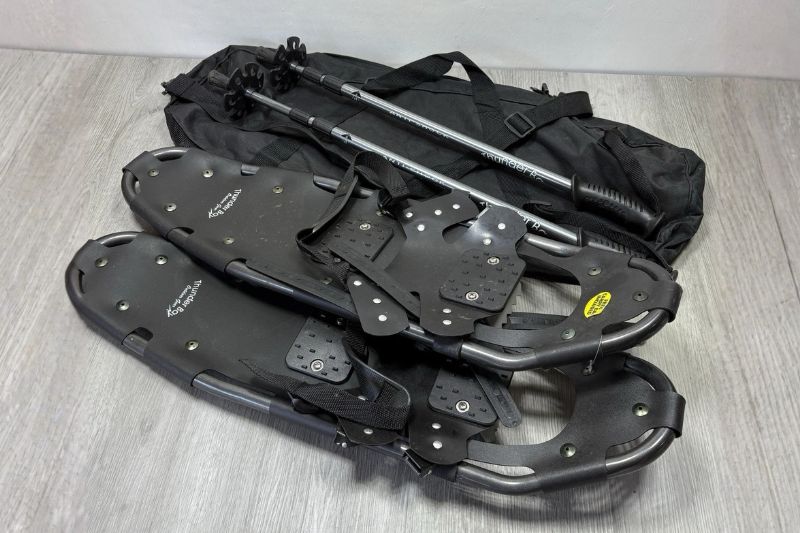7 Best Snowshoes For Winter Hiking & Mountaineering A pair of black snowshoes and two trekking poles placed on a gray wooden floor, beside a black carrying bag, conveying readiness for a snowy adventure.