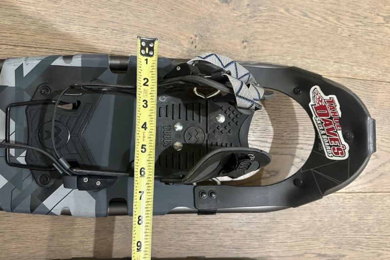 7 Best Snowshoes For Winter Hiking & Mountaineering A snowshoe on wooden floor with a yellow measuring tape showing its length at approximately 27 inches. The snowshoe has a black frame and camouflage pattern.