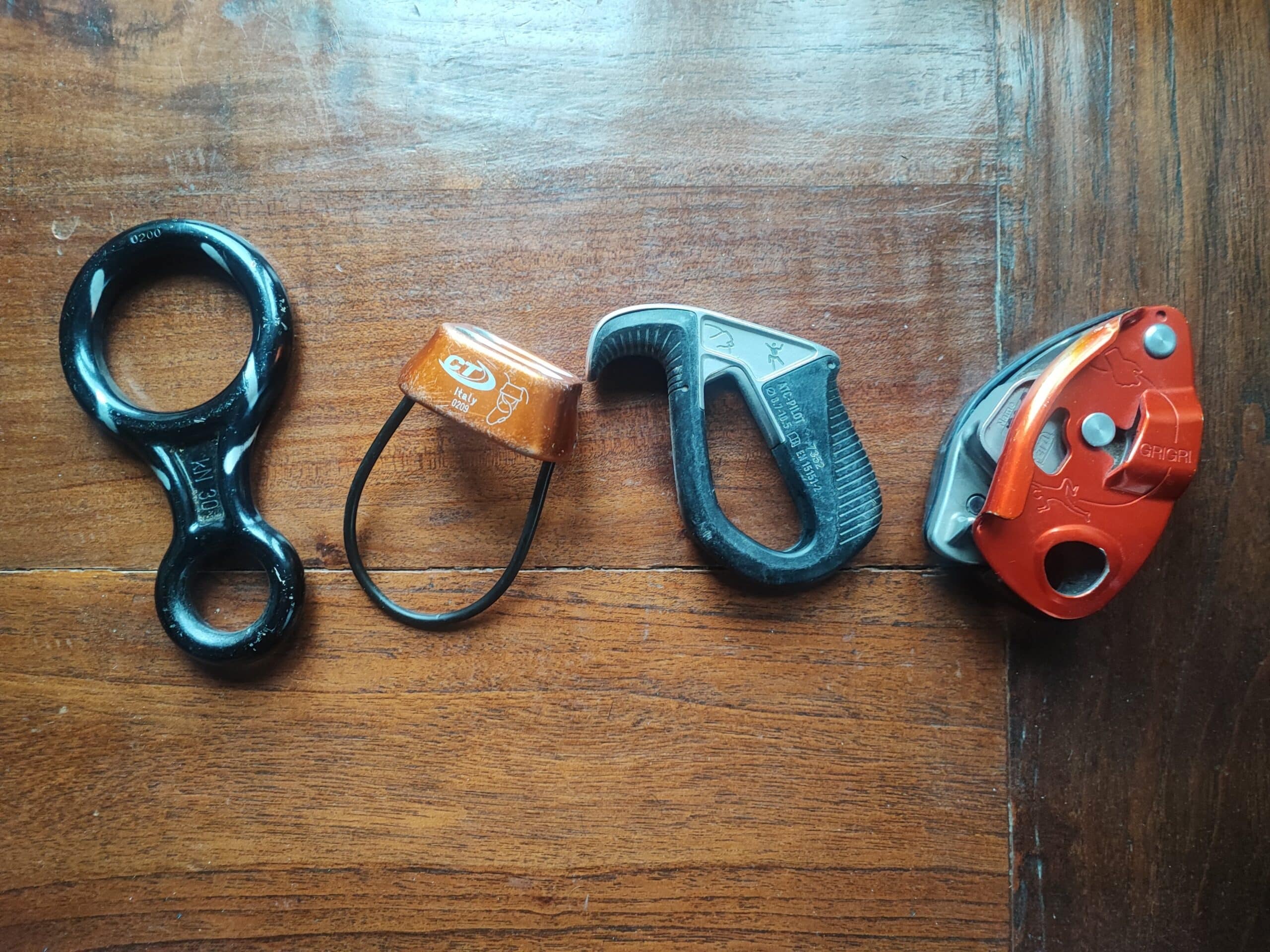 Essential Rope Skills & Knots for Mountaineering Belay Devices on a wooden surface, ranked from left to right from manual to (almost entirely) automatic. The models from left to right are: an eight, an original ATC, an ATC Pilot, a GriGri.