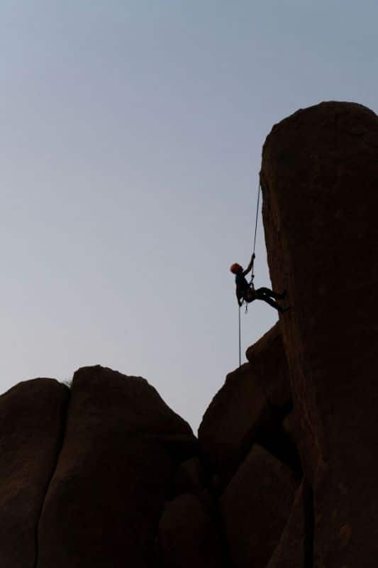 Essential Rope Skills & Knots for Mountaineering Climber rappelling during dusk. The rock and climbing appear to be black against a dim blue sky.