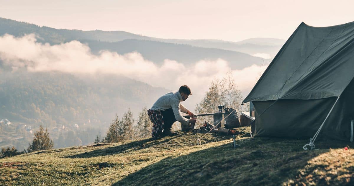 How to Turn Wild Camping Into a Glamping Experience A camper is assembling a tent on a hillside, with a scenic view of nature in the background.