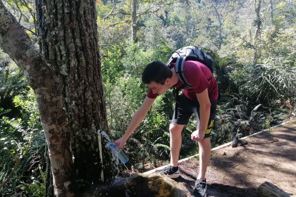 2026 Ultralight Gear Trends: The End of "Stupid Light"? A hiker bends down to fill a water bottle from a piped spring beside the trail.