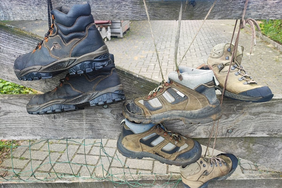 2026 Ultralight Gear Trends: The End of "Stupid Light"? Several pairs of well-worn hiking boots and lighter trail shoes, from stiff leather boots to suede-and-mesh models, drying on a wooden fence after use.