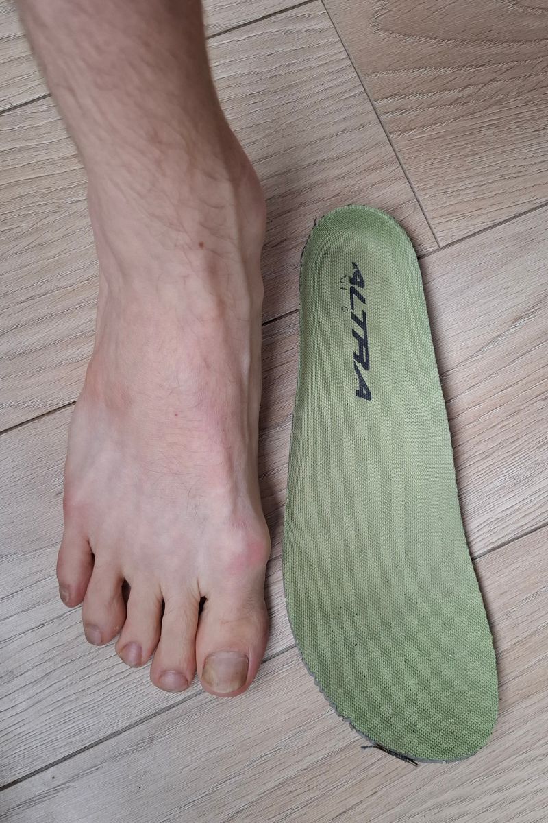 Hiking Terms: The Complete Trail & Gear Glossary. Bare foot next to a foot-shaped Altra insole on a wooden floor, the wide toe box outline matching the natural spread of the toes