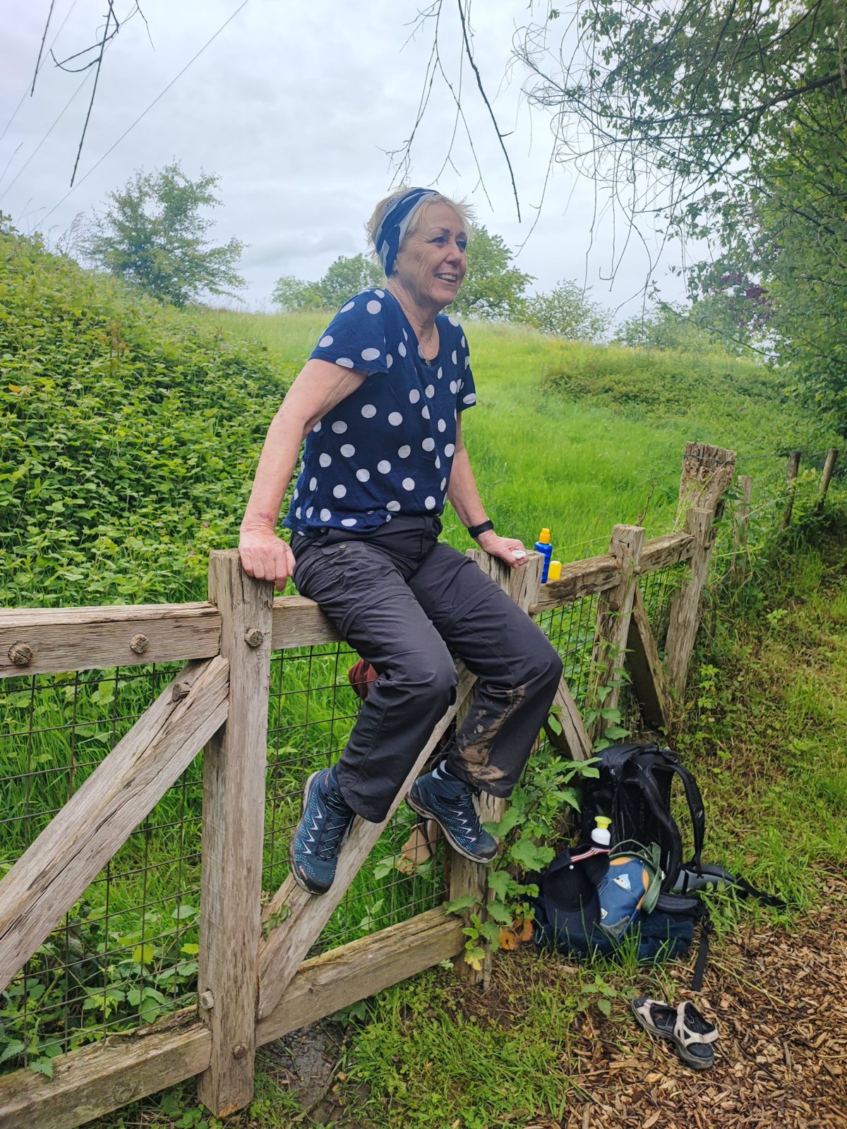 I Walked Four Days of the Pieterpad Without Booking a Single Bed Woman in a blue polka-dot top sitting on a wooden gate, laughing, with a rucksack propped against the post below.