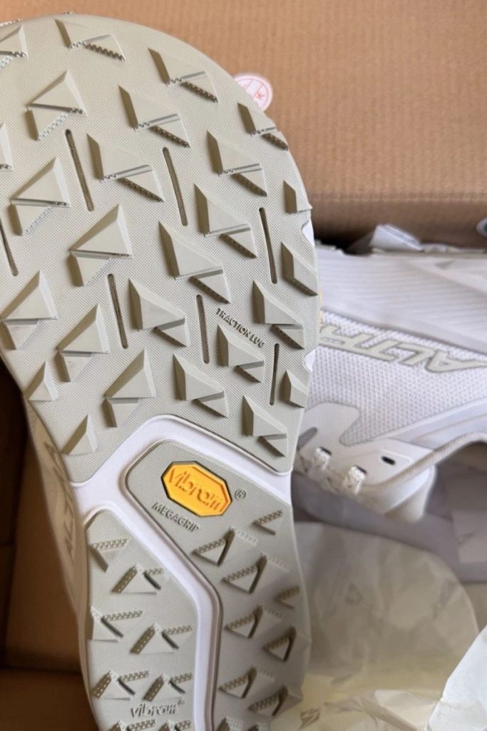 Close-up of the Altra Experience Wild 3+ sole with angular tread and yellow Vibram badge, above a matching shoe in its box.