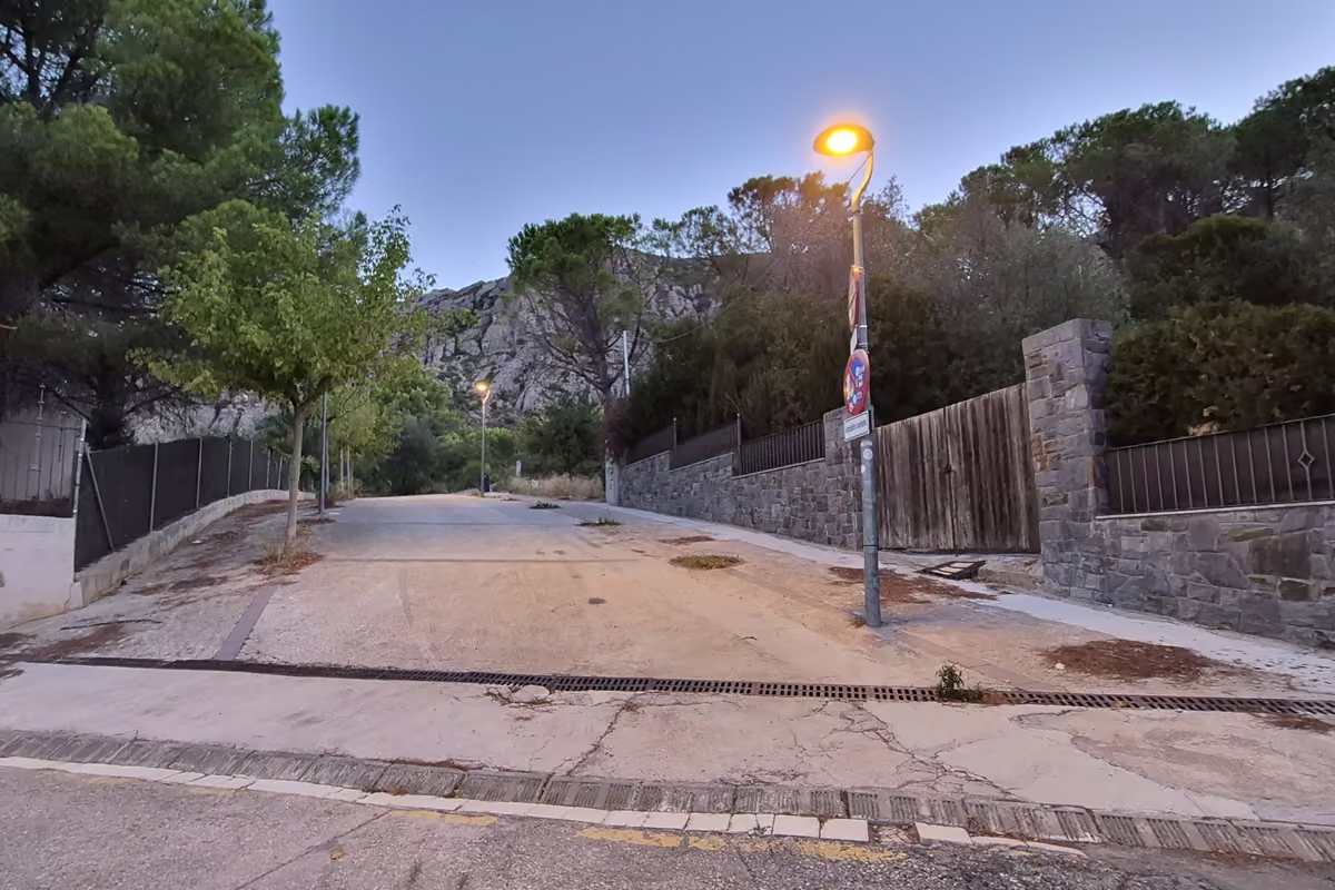 Quiet residential street at El Pujolet in Collbató at dawn, a street lamp still lit and the lower rocks of Montserrat behind the pines.