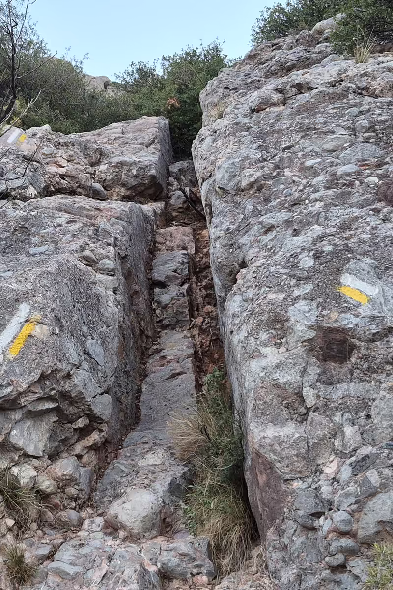 Narrow rock gully between conglomerate boulders on the Drecera de Fra Garí, marked with yellow and white PR waymarks.