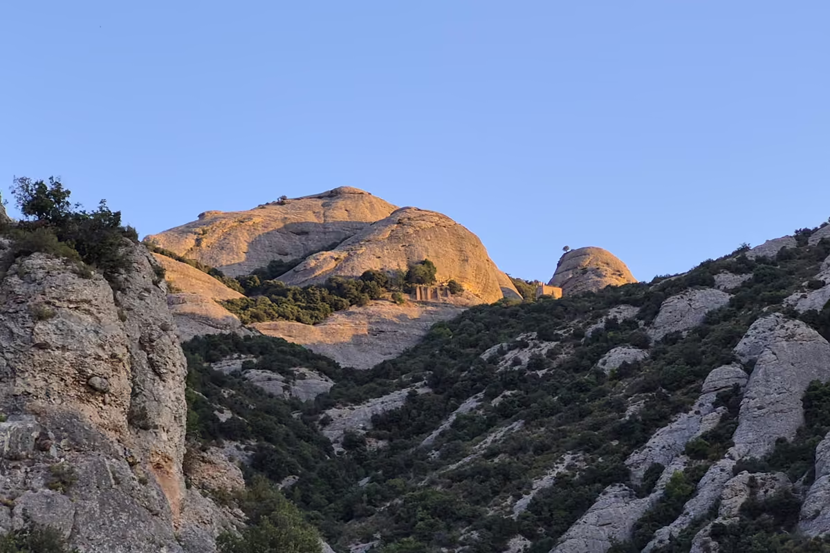 Sunlit rock domes of the Serra de Sant Joan above a wooded valley, with the hermitages of Sant Joan and Sant Onofre on the ridge in the distance.