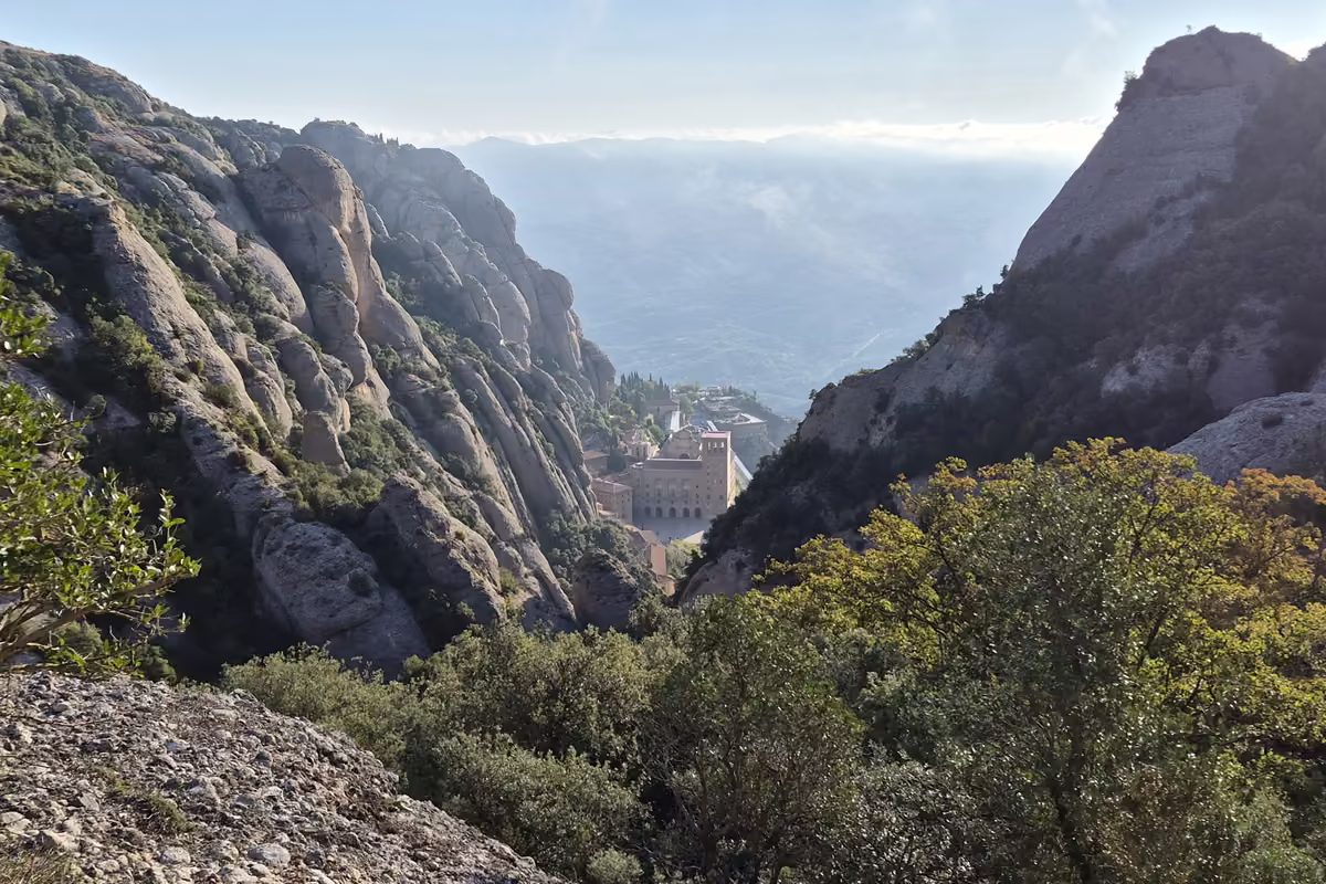 Montserrat monastery far below, framed by two steep conglomerate walls, seen from the wooded PR-C 19 path in the morning haze.