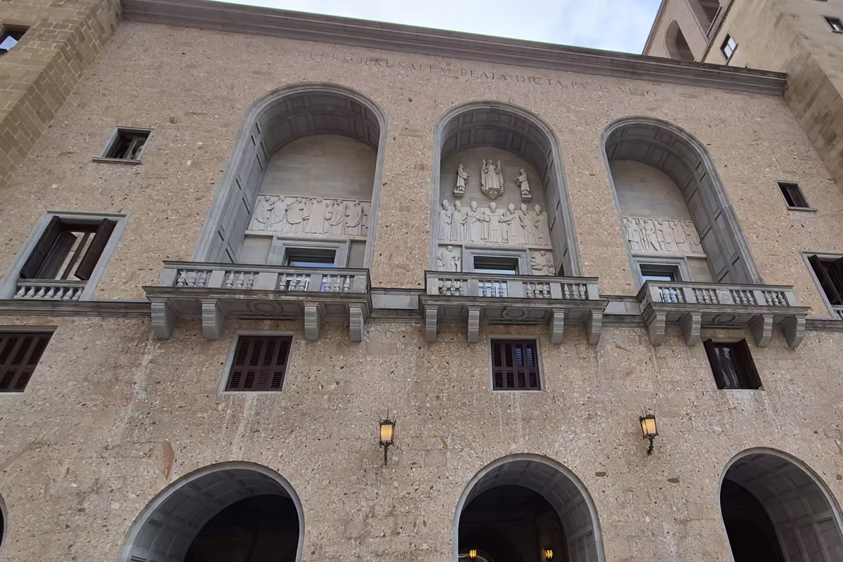 The stone façade of the Benedictine monastery of Santa Maria de Montserrat seen from below, with three arched niches holding relief sculptures.