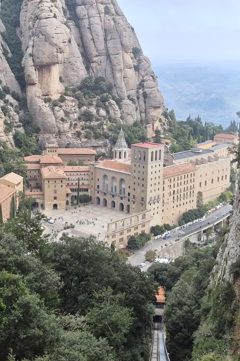 Montserrat monastery seen from high above between sheer cliffs, with the Sant Joan funicular track dropping steeply below.