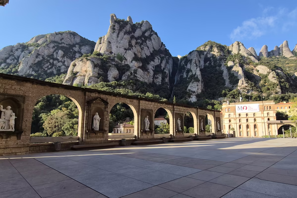 The wide paved Plaça de Santa Maria at Montserrat with a stone arcade of statues and the rock needles of the massif behind.