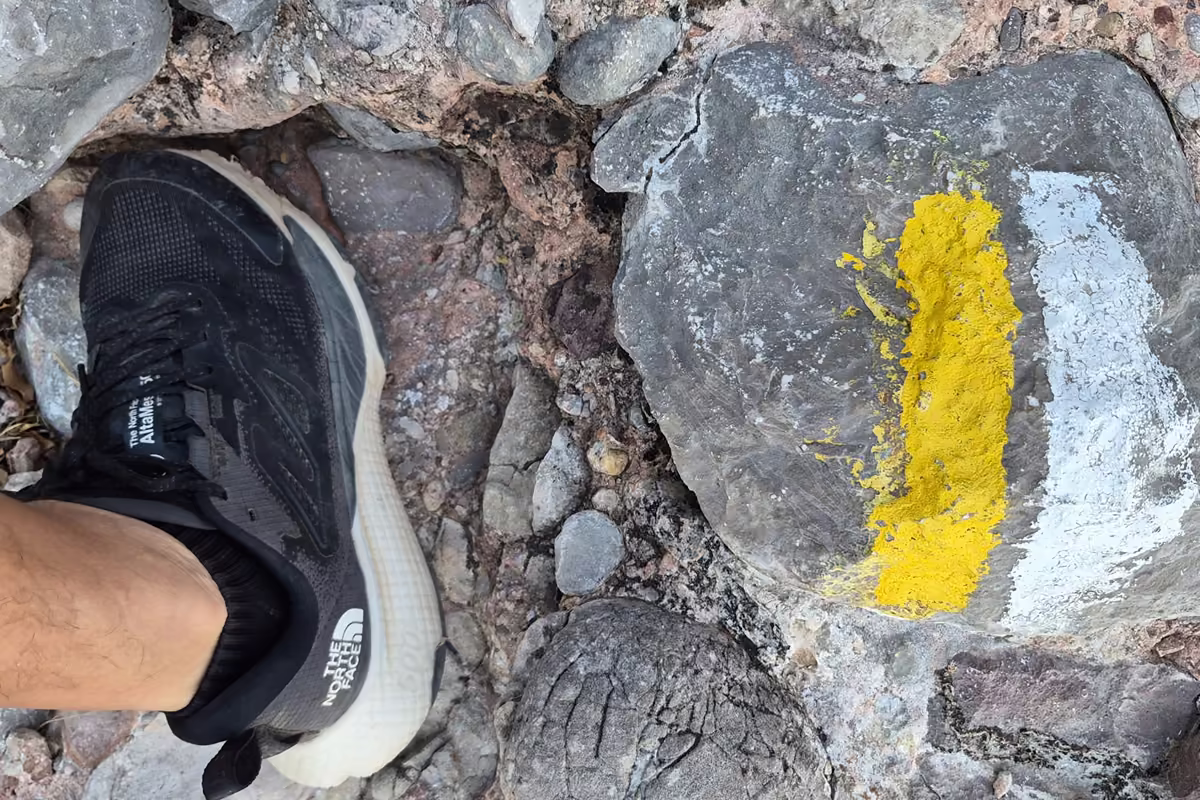 A black The North Face Altamesa 500 trail shoe on loose conglomerate stones next to a yellow and white PR waymark.