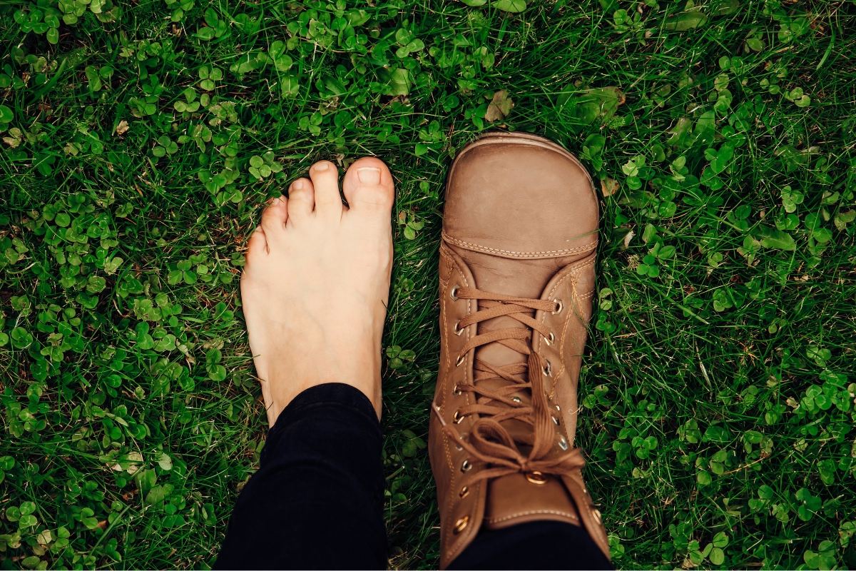 Are Barefoot Shoes Good for You? 5 Clinicians Weigh In (2026) Top-down view of a bare foot next to a brown leather barefoot boot on clover, with a toe box as wide as the foot