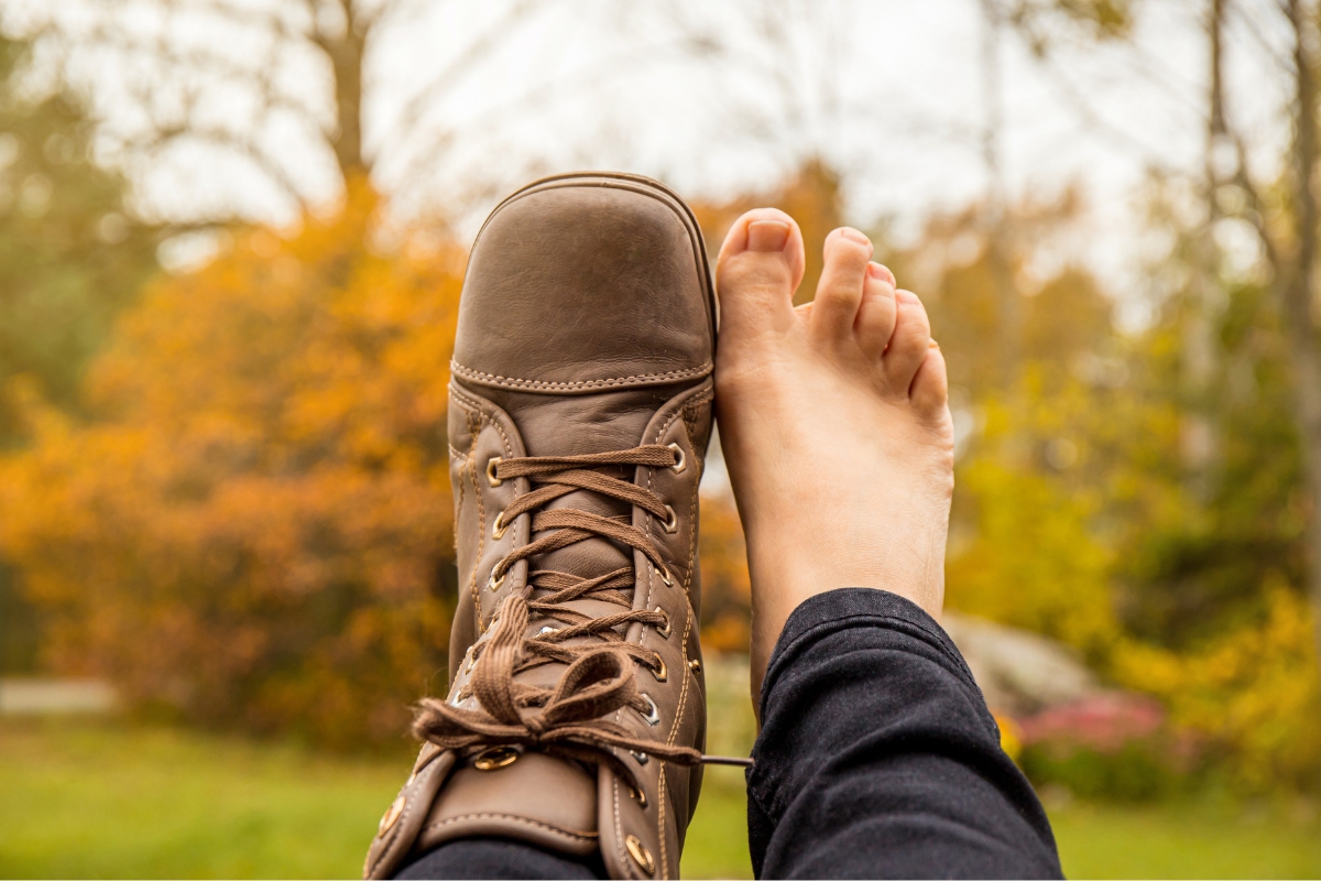 Are Barefoot Shoes Good for You? 5 Clinicians Weigh In (2026) Brown leather barefoot boot held up next to a bare foot against autumn trees, with a toe box matching the foot’s width