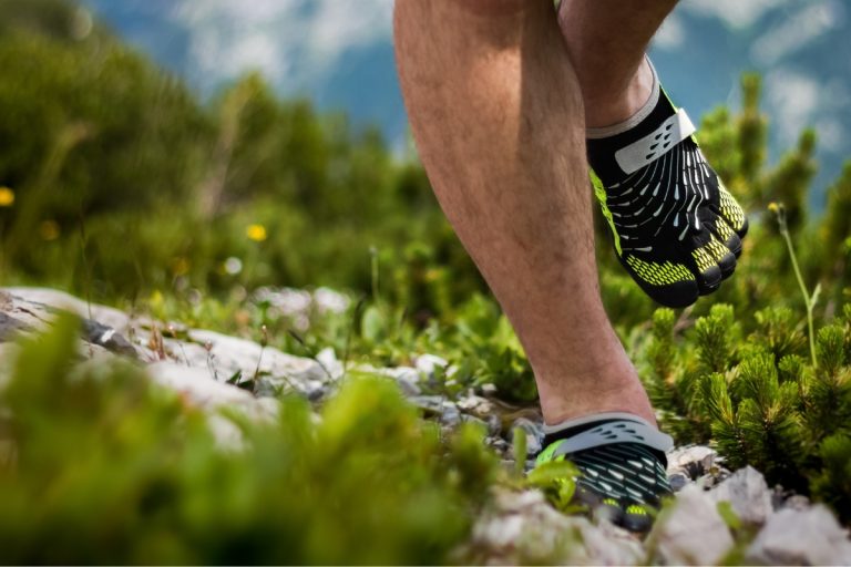 Are Barefoot Shoes Good for You? 5 Clinicians Weigh In (2026) Hiker walking a rocky mountain trail in black and yellow five-toe barefoot shoes, with low pines and peaks behind