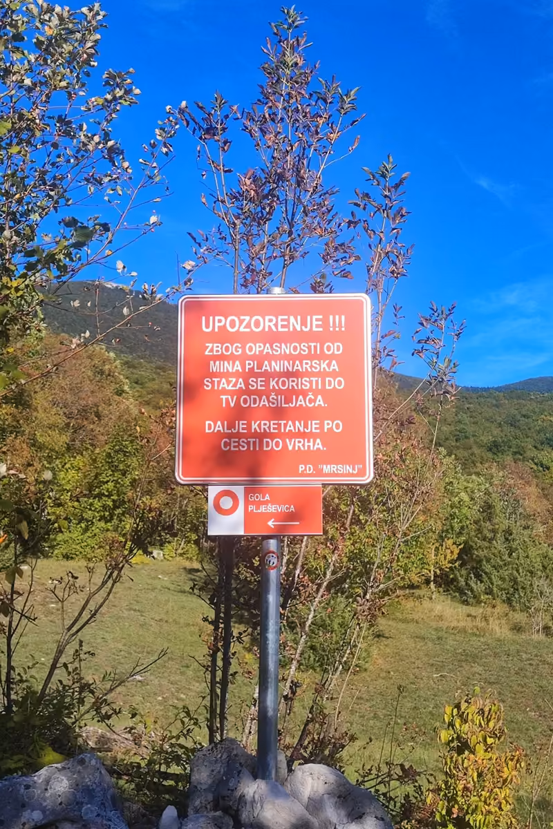 Red Croatian warning sign from the hiking club PD Mrsinj about the danger of mines, above a red and white trail marker pointing left to Gola Plješevica.