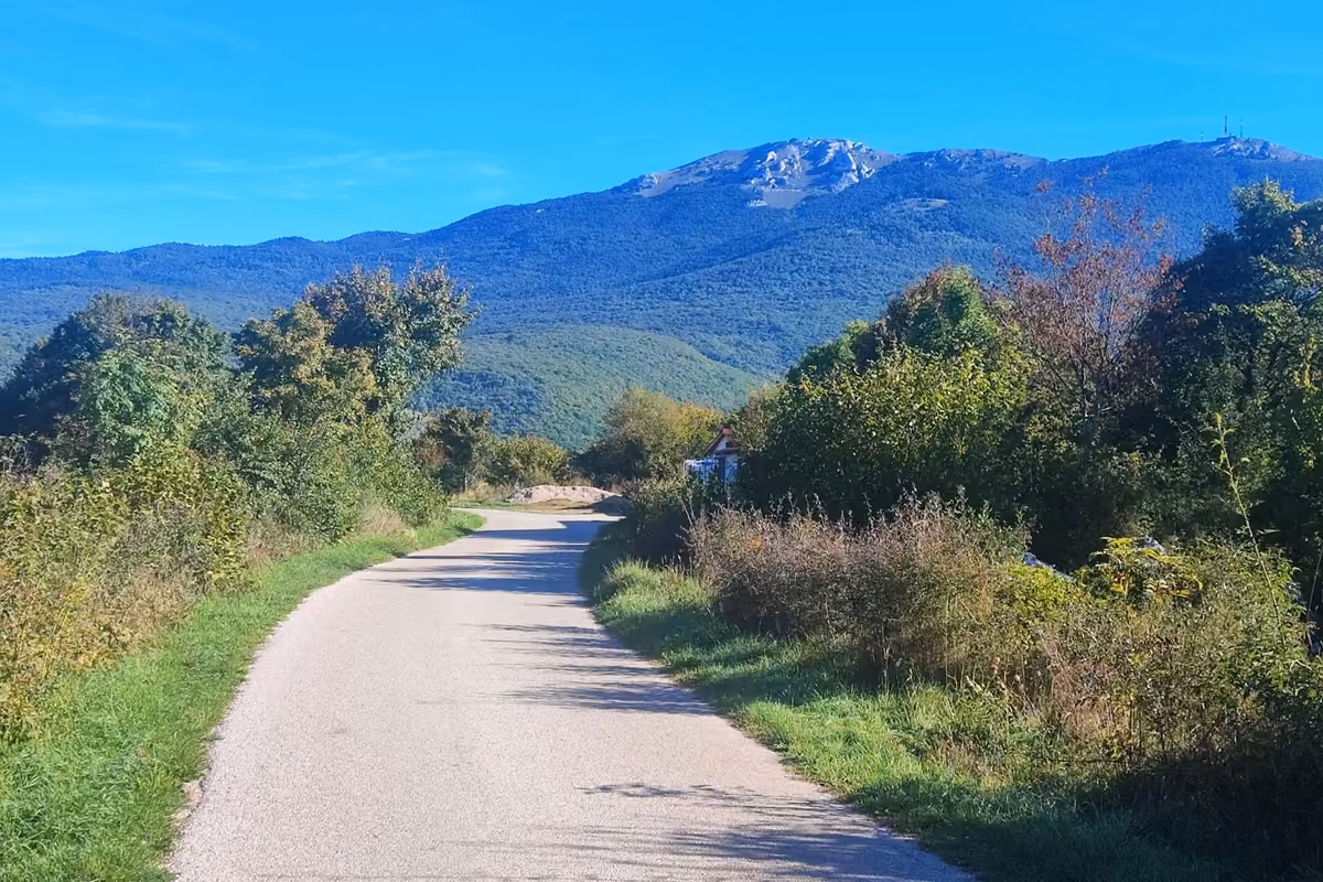 Quiet paved road between green bushes outside Korenica, with the rocky summit of Gola Plješivica and its antennas rising ahead under a clear blue sky.