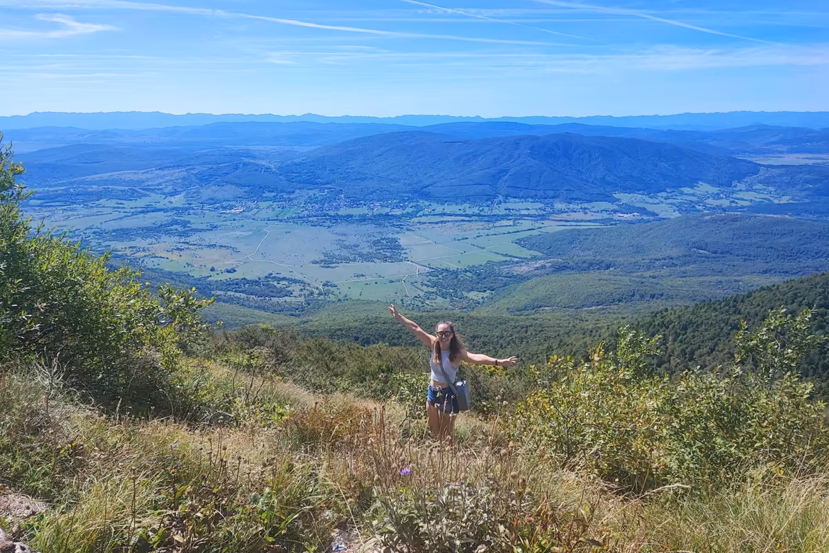 A hiker with arms spread wide on a grassy slope high above a broad green valley of fields, villages and forested hills.