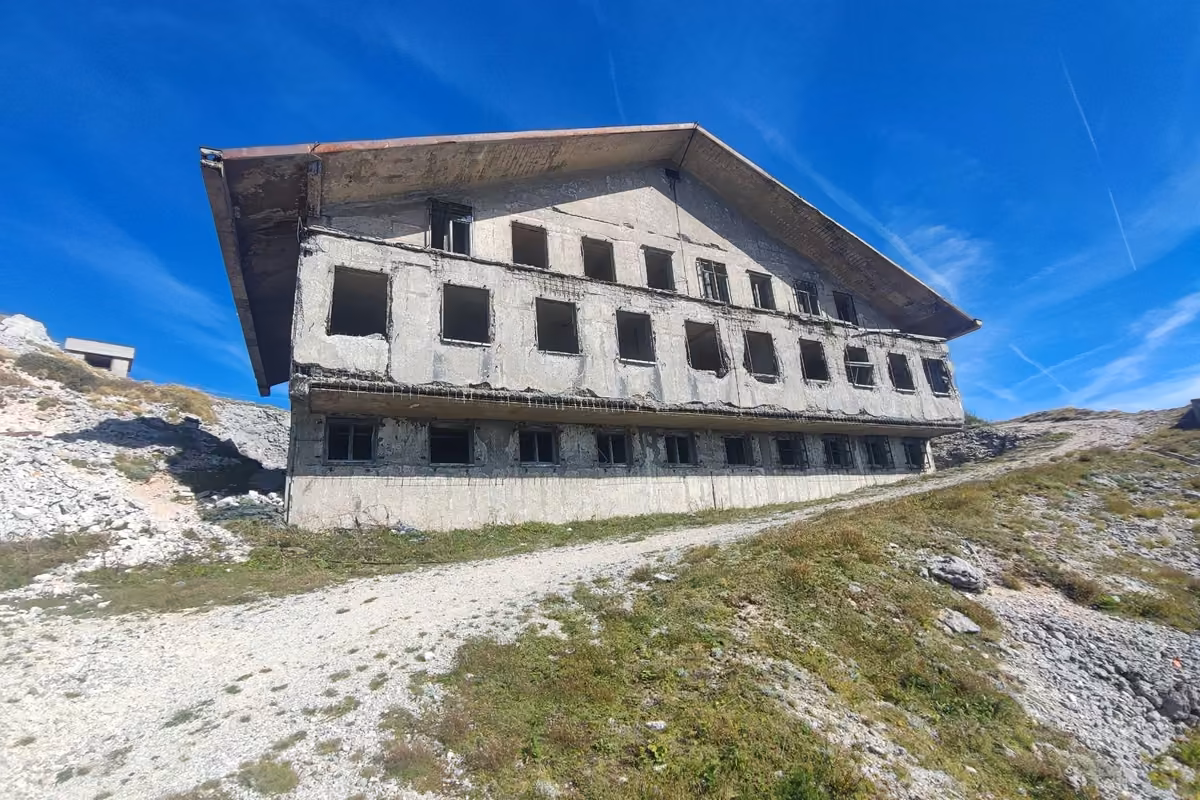Large abandoned concrete building with empty window frames and a rusted roof on the bare summit of Gola Plješivica, under a deep blue sky.