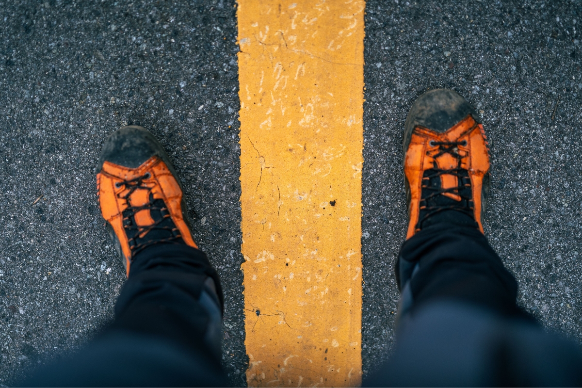 Are Barefoot Shoes Good for You? 5 Clinicians Weigh In (2026) Top-down view of feet in orange and black hiking shoes on either side of a yellow line on an asphalt road