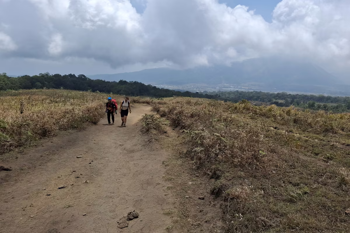 Two hikers walking up a wide, dusty dirt track through dry golden grassland under a cloudy sky, with forested hills behind them.