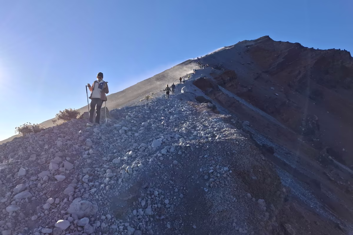 A hiker with trekking poles on a steep ridge of loose grey volcanic scree, with more hikers climbing toward the summit behind him against a bright morning sky.