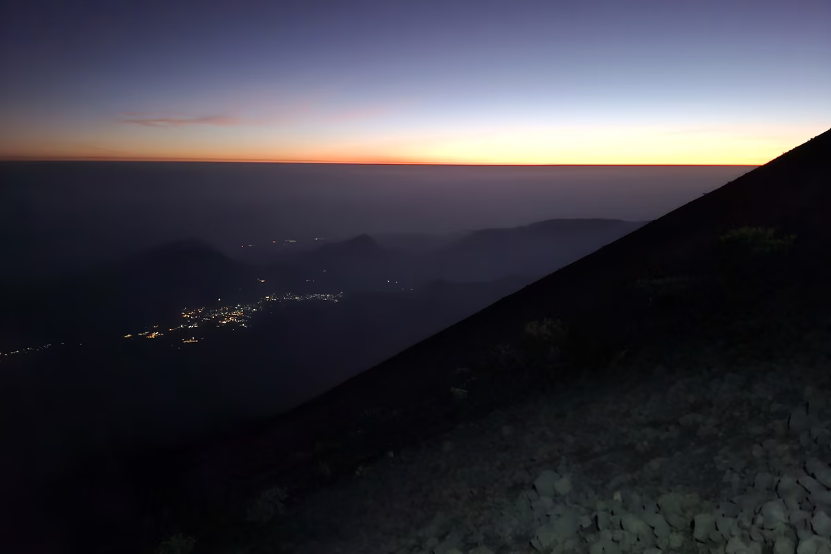 An orange band of light on the horizon above dark clouds, with village lights still glowing far below the dark flank of the volcano.