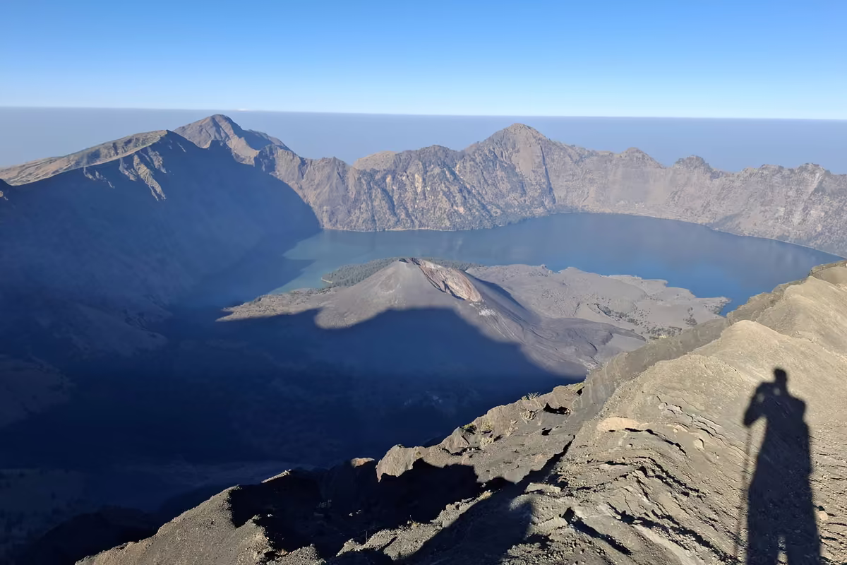 The Segara Anak crater lake and the dark Barujari cone inside the Rinjani caldera, with the shadow of a hiker holding a trekking pole on the slope in front.