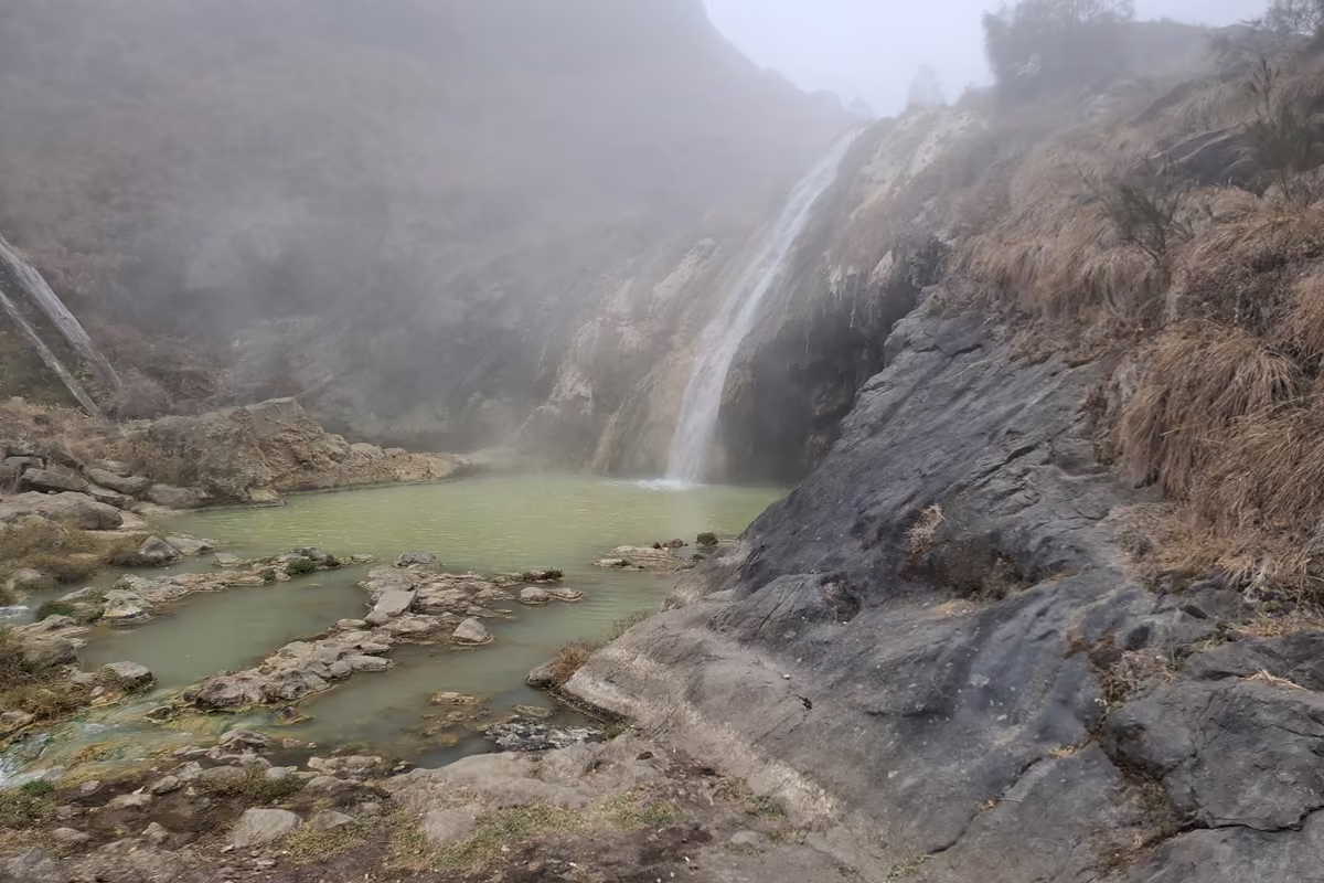 Milky green pools of a hot spring among grey rocks at the foot of a small waterfall, with steam and mist in the air.