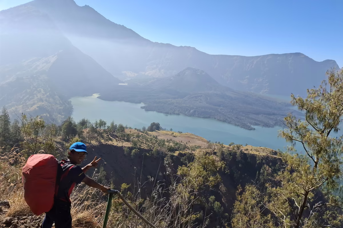A porter with a large red pack holding a rope railing on a steep path, with the crater lake and the volcano in the hazy background.