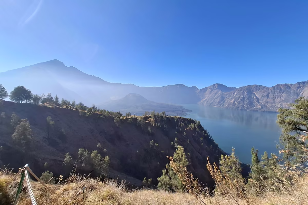 Wide view over a deep blue crater lake inside steep caldera walls, with the summit of Rinjani on the left and a rope railing in the foreground.